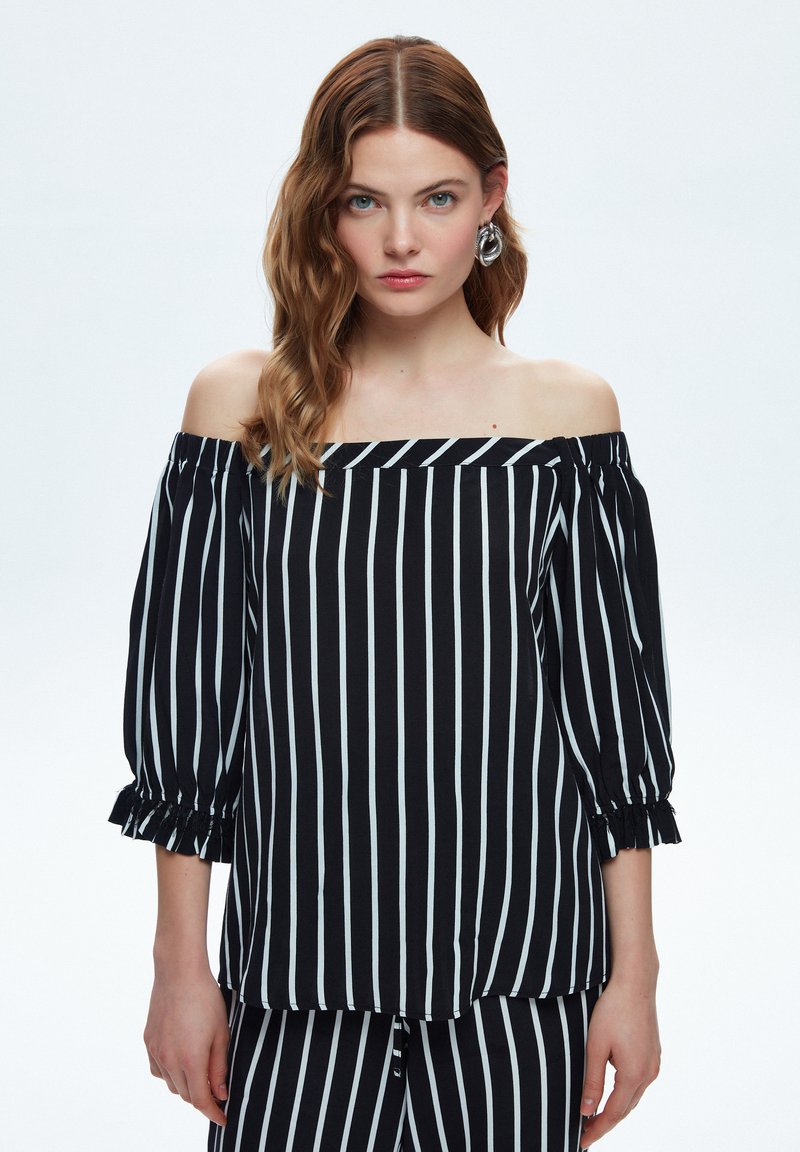Sort og hvid stribet top med off-shoulder udskæring og klokkeærmer. Har volanger i manchetten og en løs pasform, lavet af letvæv.