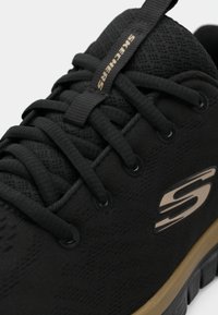 Schwarzer Sportschuh mit strukturiertem Obermaterial, Schnürung, einem goldenen Logo und einer gepolsterten Sohle mit einem kontrastierenden tanfarbigen Akzent.