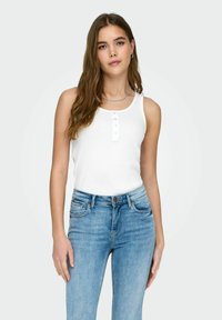 Jeune femme aux longs cheveux bruns portant un haut blanc sans manches boutonné et un jean bleu clair sur un fond uni.