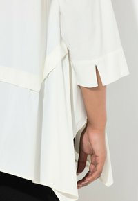 Mano accanto a una camicia bianca con manica larga e dettaglio a spacco, che mostra il tessuto a strati e una vestibilità ampia.