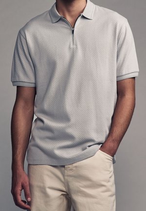 Polo gris clair avec un motif texturé, manches courtes et col à demi-fermeture éclair ; associé à un pantalon de couleur crème.