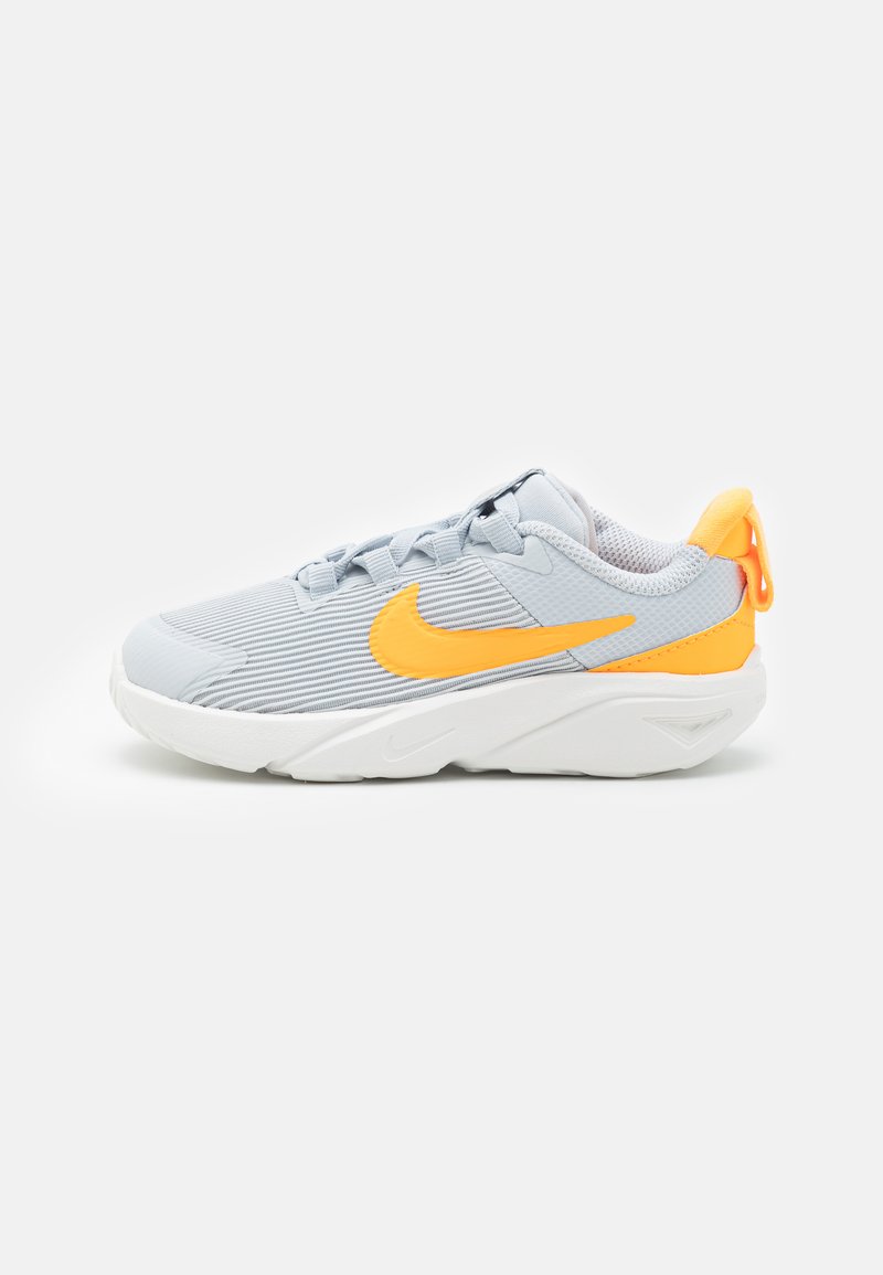 Chaussure de sport gris clair avec texture côtelée, accents orange, semelle blanche et une languette au talon pour un enfilage facile.