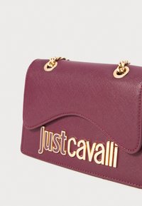 Ljubičasta kožna torba s teksturom, sa zlatnom lancem i velikim zlatnim logotipom "Just Cavalli" na prednjem klapnu.