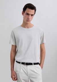 Giovane vestito con una semplice maglietta grigia chiara infilata in un pantalone bianco con una cintura nera, in piedi contro uno sfondo chiaro e uniforme.