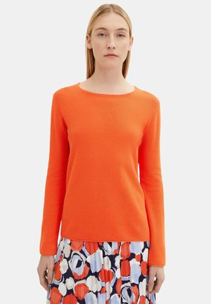 Helles orangefarbener Pullover mit langen Ärmeln, gerippter Textur und rundem Ausschnitt, kombiniert mit einem mehrfarbigen gemusterten Rock.