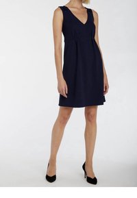 Vrouw in een mouwloze marineblauwe jurk tot de knie met een V-hals en zwarte hoge hakken, staande voor een witte achtergrond.