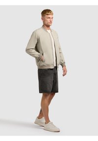 Homme debout, portant une veste bomber beige clair, un t-shirt blanc, un short en jean noir et des baskets blanches, sur un fond clair uni.