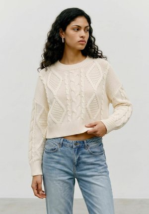 Femme aux cheveux bouclés portant un pull en maille torsadée couleur crème et un jean bleu clair sur un fond uni et clair.