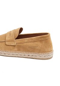 Zapato sin cordones de ante color tostado con suela de yute trenzado y una pequeña solapa decorativa. Presenta una marca en relieve en el lateral. Textura lisa en general.