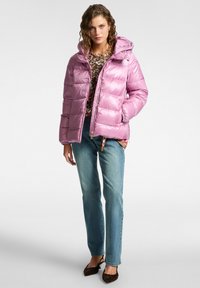 Kvinna som bär en glansig rosa puffjacka över en leopardmönstrad topp, blå jeans och mörka spetsiga klackar, stående mot en enkel bakgrund.