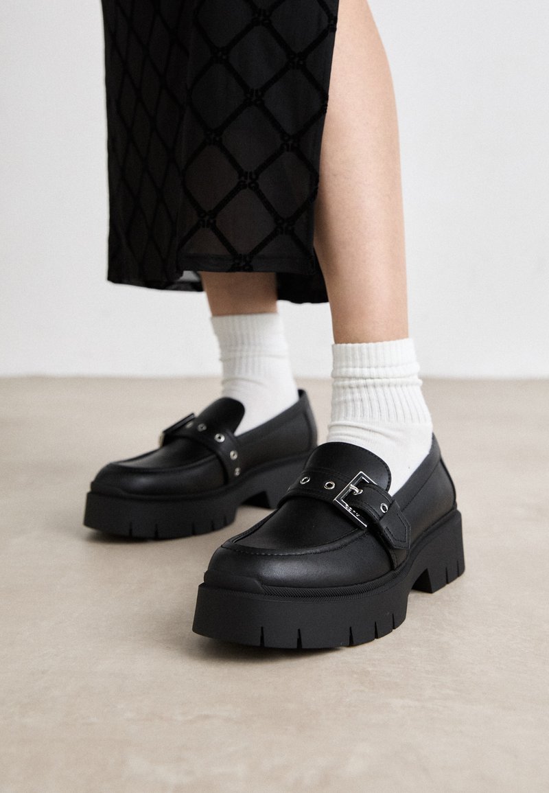 Black platform loafers with a smooth leather finish, featuring a buckle detail and stud accents, paired with white ribbed socks.