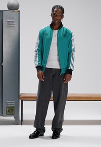 Turkis glidelåsjakke med hvite striper, svarte mansjetter; hvit skjorte under; grå vide bukser; svarte sko; garderobeskap og benk i bakgrunnen.