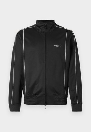 Chaqueta negra con cremallera, hecha de una tela suave, con rayas laterales blancas, puños acanalados y un cuello alzado. Logotipo en la parte frontal.