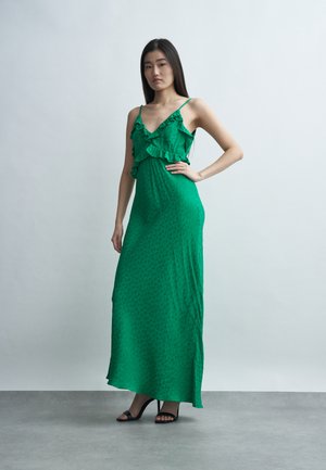 Frau steht mit einer Hand in der Hüfte, trägt ein ärmelloses grünes Maxikleid mit Rüschen und schwarze High-Heel-Sandalen vor einfachem Hintergrund.