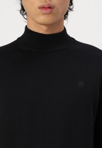 Maglione a collo alto nero realizzato in morbido tessuto a maglia, con rifiniture a coste e un piccolo logo impresso sul lato sinistro.