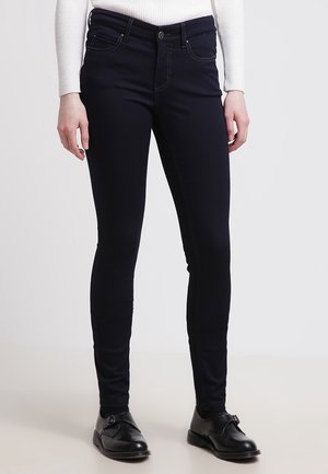 Personne portant un jean skinny noir, un top blanc à manches longues et des chaussures en cuir noir, se tenant devant un fond blanc uni.