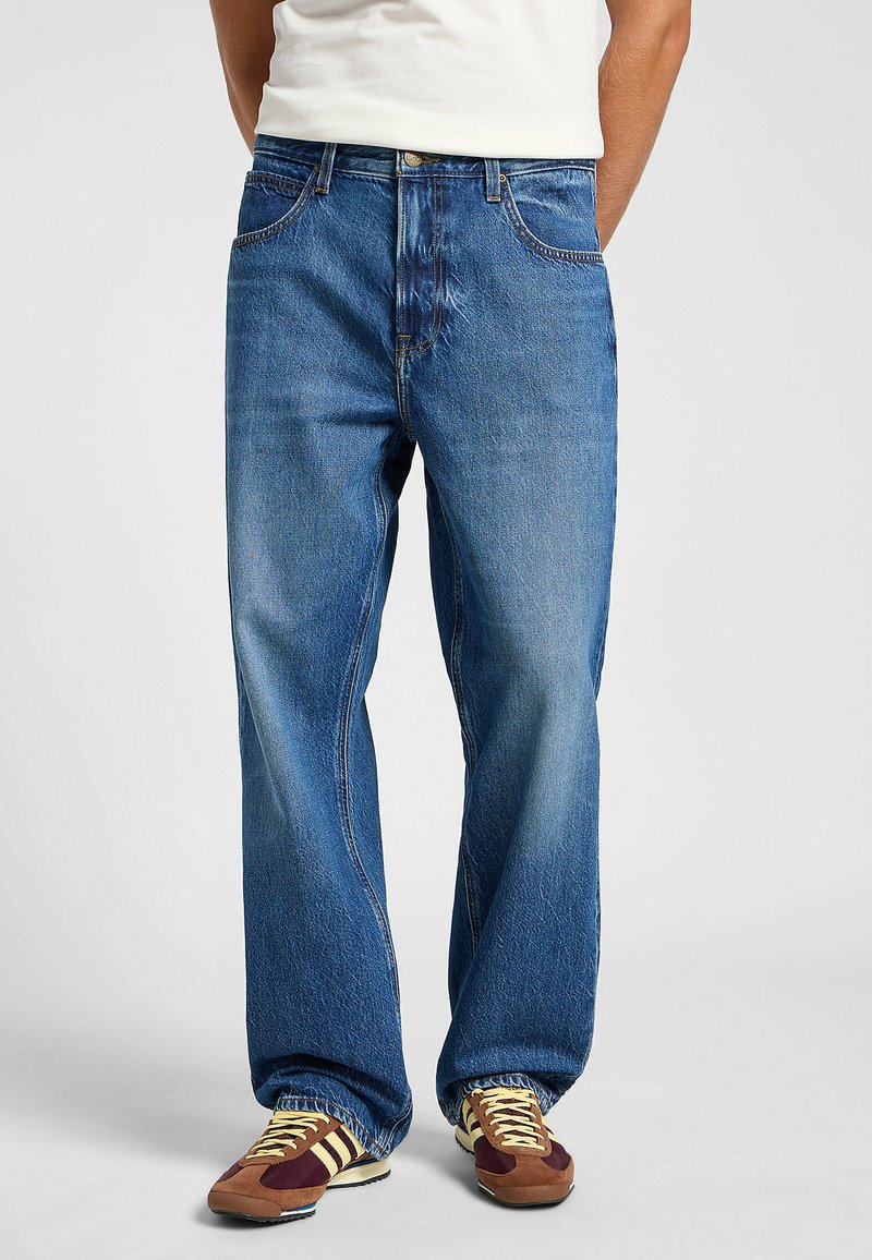 Jeans de mezclilla azul con un corte relajado, que cuentan con cinco bolsillos, trabillas para el cinturón y un acabado desgastado. Combinados con zapatillas retro marrones y amarillas.