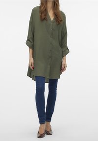 Camicia tunica verde oliva con scollo a v, chiusura a bottoni e maniche arrotolate. Indossata sopra jeans skinny blu e scarpe marroni. Texture morbida.