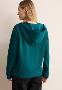 Teal hooded sweatshirt i en texturerad tygkvalitet, ribbade manschetter och en avslappnad passform. Har en känguruficka och subtil logga på ärmen.