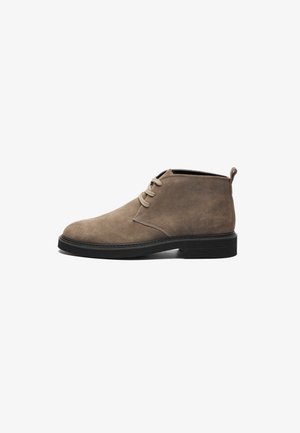 Botas de tornozelo em camurça castanho claro, com bico redondo, atacadores à frente e uma sola de borracha texturizada preta. Laço na parte de trás para fácil colocação.