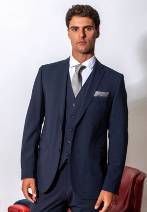 Ein Mann trägt einen dunkelblauen dreiteiligen Anzug mit silberner Krawatte und Einstecktuch, steht vor weißem Hintergrund neben einem roten Stuhl.