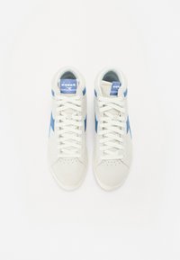 Des baskets montantes blanches avec un logo et des accents bleus, fabriquées à partir d'un mélange de matériaux lisses et en daim, avec une boîte à orteils perforée.
