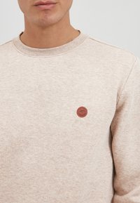 Beige sweatshirt med rund halsudskæring, tekstureret stof, med et lille brun cirkulært logo på venstre brystområde.