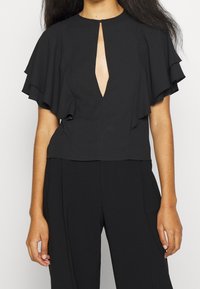 Donna che indossa una blusa nera con maniche a strati e a volants, e una fessura a chiave sul davanti, abbinata a pantaloni neri a vita alta.