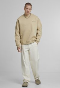 Bežová bavlnená oversize mikina s okrúhlym výstrihom a logom na ľavej hrudi, skombinovaná s bielymi voľnými nohavicami a svetlohnedými slip-on topánkami.