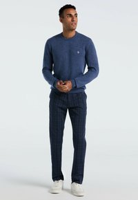 Donkerblauwe gebreide trui met een ronde hals, gecombineerd met donkergebleekte geruite broeken en witte sneakers, met een subtiel logo op de borst.