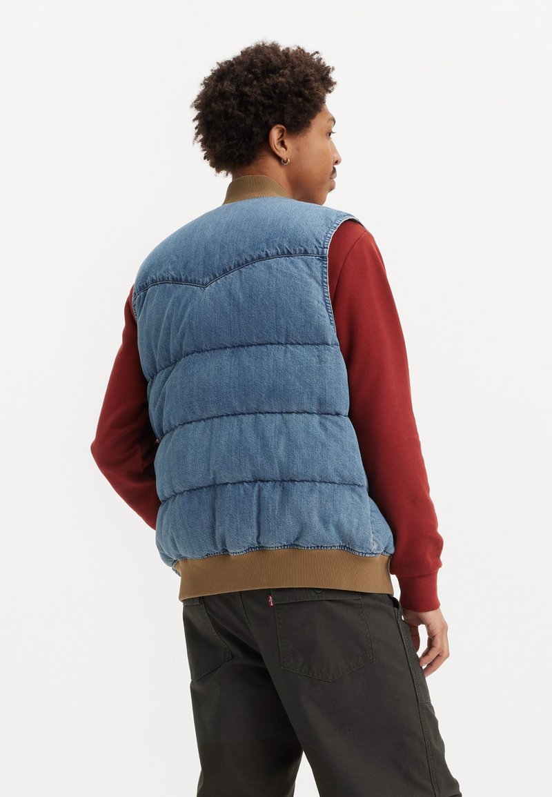 Gilet en jean, bleu clair avec des sections matelassées, associé à une chemise à manches longues bordeaux et un pantalon foncé, avec un ourlet côtelé.
