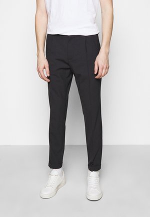 Uomo con pantaloni aderenti neri, calzini bianchi, scarpe da ginnastica bianche e una maglietta bianca semplice, in piedi davanti a uno sfondo bianco.