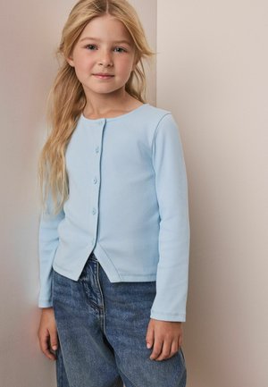 Blond dziecko ubrane w jasnoniebieski rozpinany sweter i luźne niebieskie dżinsy stoi przy beżowej ścianie.