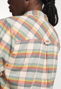 Checkered shirt featuring a light blue, beige, and orange color palette. It has a collar, pleat detail, and a looped fabric accent at the back.