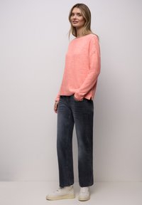 Korallenroter Pullover mit strukturierter Strickoptik und Knöpfen an den Schultern. Kombiniert mit dunkelgrauen Jeans und weißen Turnschuhen.