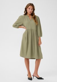 Olivgrünes, knielanges Kleid mit strukturiertem Finish, 3/4 langen Puffärmeln, einem gesplitteten Ausschnitt und einem gerafften Bund. Kombiniert mit schwarzen High-Heel-Mules.