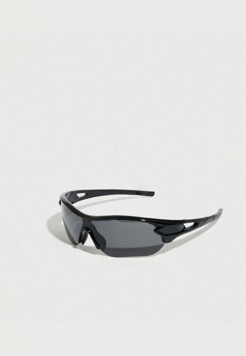 Μαύρα σπορ γυαλιά ηλίου τύπου wraparound με σκούρους φακούς και καμπυλωτό σκελετό, τοποθετημένα σε ανοιχτόχρωμη επιφάνεια.