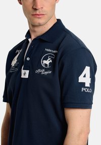 Polo de color azul marino hecho de una tela texturizada, con un color blanco en el cuello, logo bordado y el número "4" en la manga.