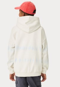 Sweat à capuche surdimensionné blanc avec un motif tie-dye bleu clair au dos, présentant une grande capuche et des poignets côtelés, associé à une casquette de baseball rouge.
