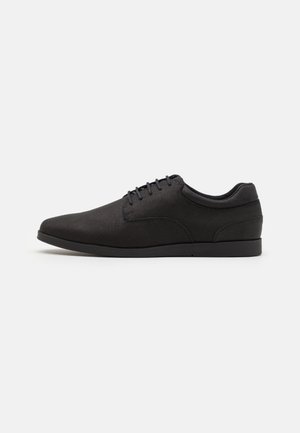 Sort lav, snørelukket dresssko med glat finish og minimalistisk design, vist i profil mod en ensfarvet baggrund.
