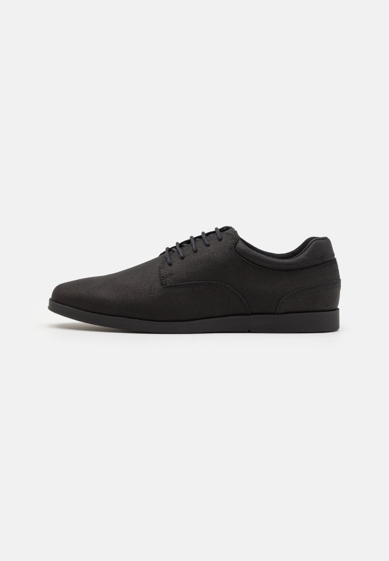Chaussure noire habillée à lacets basse avec une finition lisse et un design minimaliste, montrée de profil sur un fond uni.