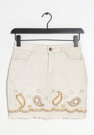 Beige denimrok met voorzakken, knoopsluiting, rafelige zoom en goud-witte paisley-borduursels langs de onderrand, hangend aan een zwarte hanger.
