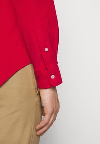 Camicia rossa a maniche lunghe con tessuto testurizzato. Presenta polsini con bottoni rosa chiaro e orlo curvo, abbinata a pantaloni color cachi.