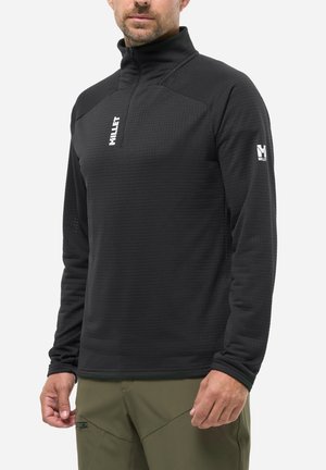 Mann trägt schwarzen Millet Fleece mit Reißverschluss und Logo auf Brust und Ärmel, kombiniert mit olivgrüner Hose, vor weißem Hintergrund.