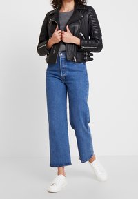 Giacca in pelle nera da motociclista con spalle rigate, abbinata a jeans blu a vita alta. Le sneakers bianche completano l'outfit.