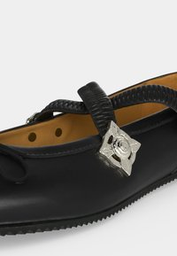 Chaussure plate en cuir noir avec bride texturée et boucle décorative argentée en forme de fleur, doublure intérieure couleur fauve et bord de semelle rainuré.