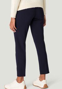 Personne portant un pantalon slim bleu marine foncé, un pull en tricot crème et des baskets blanche cassé avec des semelles en caoutchouc, debout devant un fond clair et uni.