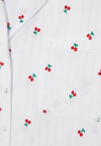 Witte blouse met rode kersborduursels, groene stelen en een borstzak. Verticale textuur is zichtbaar in de stof.
