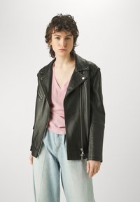 Giacca da moto in pelle nera con chiusura con zip e maniche testurizzate, indossata sopra una maglietta rosa chiaro e pantaloni di jeans azzurri.