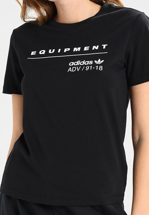 Kobieta w czarnej koszulce Adidas z nadrukiem "EQUIPMENT" i "adidas ADV / 91-18" w kolorze białym na piersi.